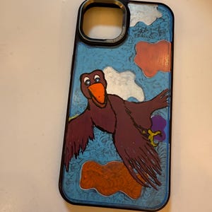 May include: A black phone case featuring a cartoon bird flying in a blue sky with white clouds and orange and purple shapes. The case has a silver ring around the camera lens with the text "100% FOCUS POWER" printed on it.