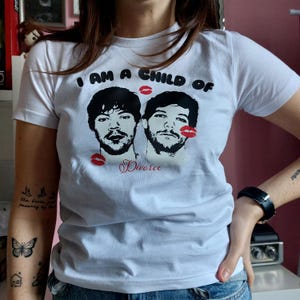 Op de afbeelding: Wit T-shirt met een grafische print van twee mannengezichten met rode lippen erboven gedrukt. De tekst "I am a child of Divorce" is onder de gezichten gedrukt.