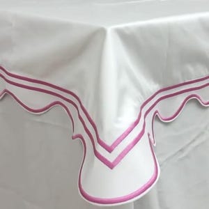 May include: White tablecloth with a scalloped edge and a double magenta border. The fabric appears to be a smooth, satin-like material. The corner of the table is visible, showcasing the design.
