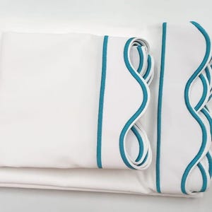 May include: White bedding with a teal scalloped trim. The bedding is folded in a stack.