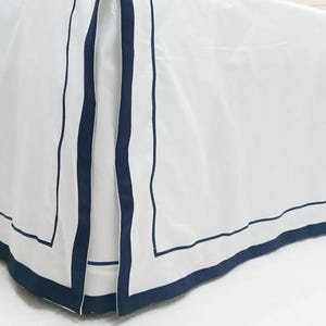 Op de afbeelding: Een witte bedrok met een donkerblauwe rand. De rand heeft twee lijnen met donkerblauwe bies.