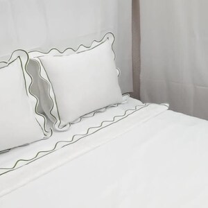 May include: White bedding set with green scalloped trim. The set includes two pillows and a fitted sheet. The pillows and sheet have a decorative green trim. The bedding is on a bed.
