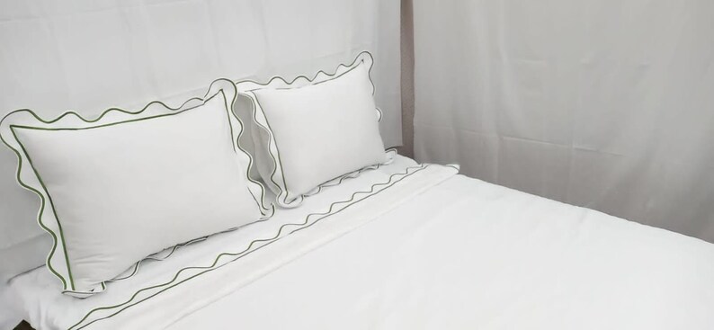 May include: White bedding set with green scalloped trim. The set includes two pillows and a fitted sheet. The pillows and sheet have a decorative green trim. The bedding is on a bed.