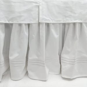 May include: White bed skirt with a ruffled edge and a simple design. The skirt has a plain white fabric with a subtle texture.