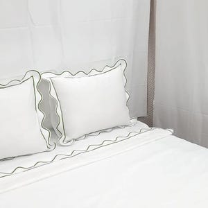 May include: White bed linens with a scalloped green trim. Two white pillows with green trim are on top of the bed. The bed is made and ready for use. The sheets and pillowcases are a solid white color.