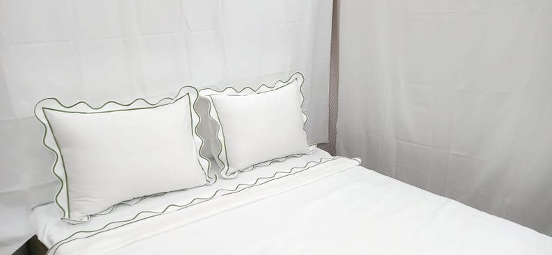 May include: White bed linens with a scalloped green trim. Two white pillows with green trim are on top of the bed. The bed is made and ready for use. The sheets and pillowcases are a solid white color.