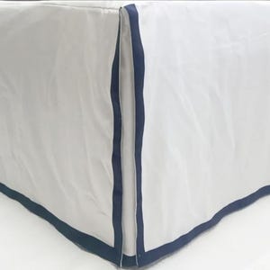 May include: White bed skirt with a navy blue trim. The skirt has a pleated design and is made of a soft, smooth fabric.
