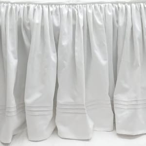 May include: White bed skirt with a ruffled edge and a simple design. The skirt has three rows of stitching along the bottom edge.