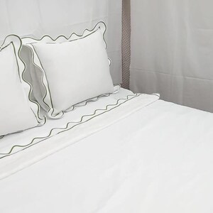 May include: White bed linens with green scalloped trim. The set includes two pillows and a fitted sheet, all with a decorative green border. The bedding is neatly arranged on a bed, creating a clean and inviting look.