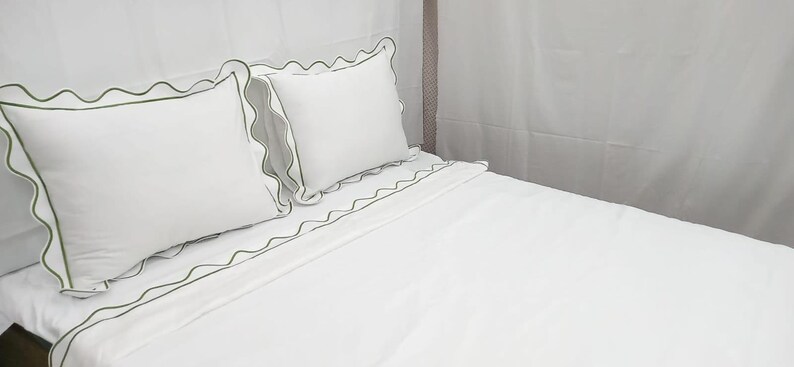 May include: White bed linens with green scalloped trim. The set includes two pillows and a fitted sheet, all with a decorative green border. The bedding is neatly arranged on a bed, creating a clean and inviting look.