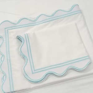 May include: Two white pillowcases with a scalloped edge and a light blue trim. The pillowcases are folded on top of each other.
