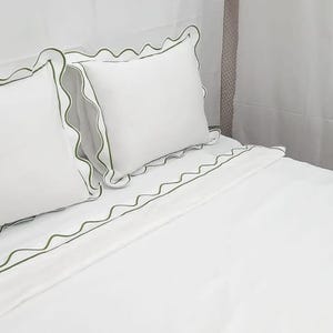 May include: White bedding set with green trim. The set includes two pillowcases and a fitted sheet. The pillowcases and the sheet have a scalloped edge with a green border. The bedding is made of a soft, smooth fabric.