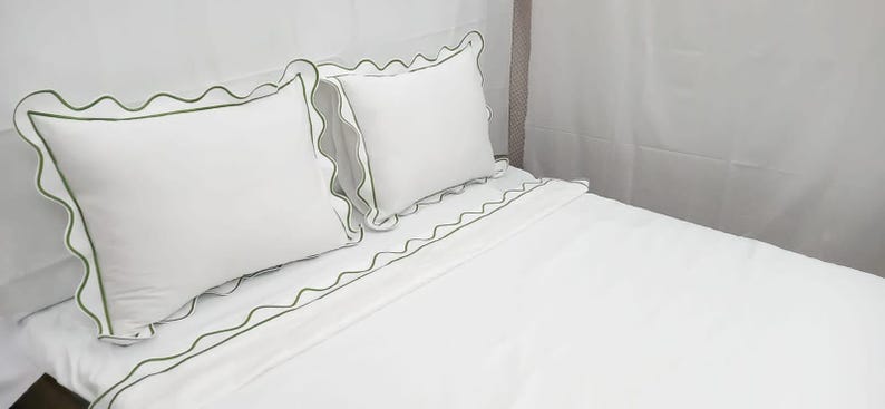 May include: White bedding set with green trim. The set includes two pillowcases and a fitted sheet. The pillowcases and the sheet have a scalloped edge with a green border. The bedding is made of a soft, smooth fabric.