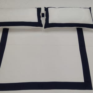 Puede incluir: Sábana blanca de cama con un borde azul marino. Dos almohadas blancas con bordes azul marino están encima de la sábana.