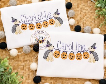 May include: Two white shirts featuring embroidered designs of two light-colored dogs wearing purple witch hats and capes. The dogs hold a string of five orange jack-o'-lantern buckets. The names Charlotte and Caroline are stitched in purple.