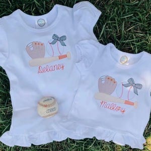 Op de afbeelding: Twee witte T-shirts met geborduurde baseball-ontwerpen. Het bovenste shirt heeft een honkbal, een knuppel, een handschoen en een strik met de naam "Delaney" eronder geborduurd. Het onderste shirt heeft een honkbal, een knuppel, een handschoen en een strik met de naam "Mallory" eronder geborduurd.