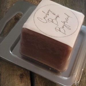 May include: A bar of soap with a two-tone design, brown at the bottom and white at the top, resting on a silver tray. The top of the soap has the words "Cowboy Boyfriend" in a circular design. The soap is on a wooden surface.