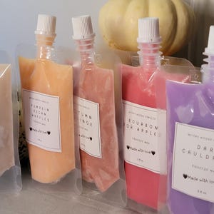 May include: Several squeeze wax pouches in shades of peach, pink, and purple. Each pouch has a white label with text like "Autumn Equinox" and "Bourbon for Apple." A decorative pumpkin is visible in the background.
