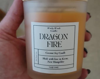 Può includere: Una candela in un barattolo di vetro smerigliato con coperchio in legno chiaro e cera arancione. L'etichetta bianca riporta la scritta Witchy Woods Candles, DRAGON FIRE, Coconut Soy Candle, Made with love in Keene, New Hampshire, e 7OZ/198G.