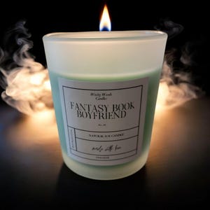 May include: A lit, frosted glass candle with a green interior and a label that reads "Fantasy Book Boyfriend." The candle is labeled "Natural Soy Candle" and is surrounded by wisps of smoke against a dark background. The candle is 283g.