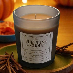 Puede incluir: Una vela de cristal esmerilado con cera de color naranja claro y una mecha. La etiqueta dice "PUMPKINS & CHOULI" y "Coconut Soy Candle". La vela está sobre un plato verde con palitos de canela, con calabazas en el fondo.