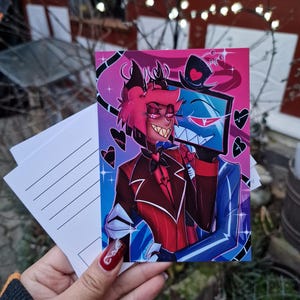 Könnte beinhalten: Eine farbenfrohe Postkarte mit zwei Zeichentrickfiguren. Eine Figur hat rotes Haar und einen roten Anzug, die andere ist blau mit Zylinder. Der Hintergrund wechselt von Pink zu Blau, mit schwarzen Herzakzenten. Mehrere leere Postkarten sind sichtbar.