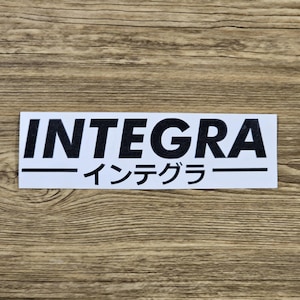 May include: A white rectangular sticker with the word "INTEGRA" in bold black letters. Below the word is a black line, and beneath that, Japanese characters. The sticker is on a wooden surface.