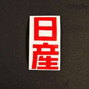 May include: A rectangular sticker with the Japanese characters for "Made in Japan" in red against a white background. The sticker is on a black surface.
