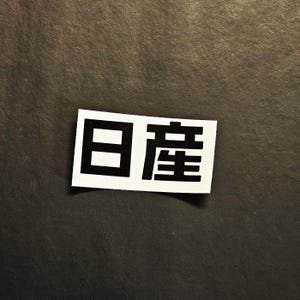 May include: A white rectangular sticker with black Japanese characters. The sticker is on a dark gray surface. The Japanese characters are in a bold, sans-serif font.