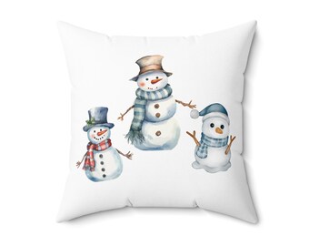 Op de afbeelding: Wit vierkant kussen met een aquarel illustratie van drie sneeuwmannen. Elke sneeuwman draagt een andere hoed en sjaal, met takkenarmen en knoopdetails. De sneeuwmannen zijn in tinten blauw, rood en bruin.