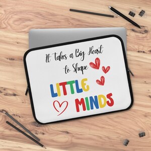 May include: White laptop sleeve with a black border, featuring the text "It takes a Big Heart to Shape LITTLE MINDS" in various colors and a heart graphic. The sleeve is on a wooden surface with pencils and a pen.