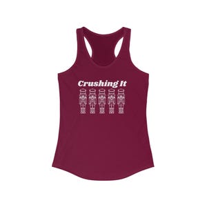 May include: A maroon tank top with a racerback design. The front features the text "Crushing It" above a row of white line-art nutcracker figures. This top is a casual wear item.