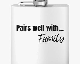 Może przedstawiać: Biała piersiówka ze srebrną nakrętką i zawiasem. Na piersiówce czarnym drukiem napisano "Pairs well with... Family". Przeznaczona do przechowywania płynów.