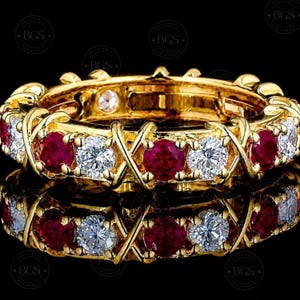May include: A yellow gold eternity ring featuring an alternating pattern of round-cut red rubies and clear white diamonds. The stones are set within a continuous gold band characterized by an X-shaped metal design between each gemstone.