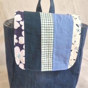 May include: A denim backpack with a patchwork flap. The flap features a floral print, dark teal, green gingham, and light blue fabric panels. The bag has a top handle and is made of blue denim.