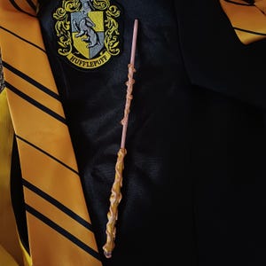 May include: A detailed wand with a light pink shaft and a textured, golden handle. The wand is set against a black robe and a yellow and black striped tie, with a Hufflepuff crest visible in the background.
