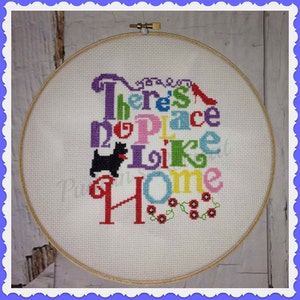 May include: Cross-stitch embroidery in a wooden hoop. The design features the phrase "There's no place like home" in various colors, with a black dog silhouette and a red high heel.