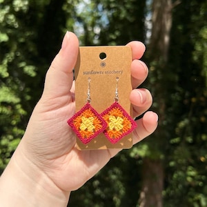 May include: Crocheted square earrings in red, orange, and yellow on a brown card with the text "sunflower stitchery".