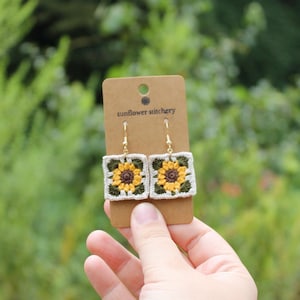 Op de afbeelding: Gehaakte zonnebloem oorbellen met goudkleurige haken. De vierkante oorbellen hebben een witte rand, groene bladeren en een geel-bruin zonnebloemmotief. De oorbellen worden gepresenteerd op een bruine kaart met de tekst "sunflower stitchery".