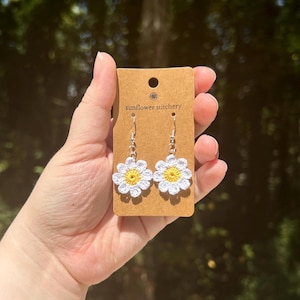 May include: A pair of crocheted daisy earrings with yellow centers and white petals. The earrings are on a brown card with the text "sunflower stitchery".