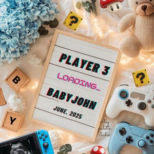May include: A white letter board with the text "PLAYER 3 LOADING... BABY JOHN JUNE. 2025" surrounded by blue hydrangeas, a teddy bear, a video game controller, and a Nintendo Switch.