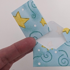May include: A small, open, blue envelope with a star and swirl pattern. The envelope has a heart-shaped closure. The design includes yellow stars and white dots. The envelope is held open, revealing a white interior.