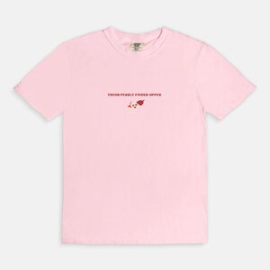May include: Pink short-sleeved t-shirt with the text "Cocoa Pebble Picker-Upper" and a small graphic of a red cocoa pebble and a scoop.