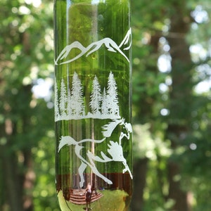 May include: A repurposed green glass bottle bird feeder with etched mountain, forest, and horse designs. The feeder has a curved, clear plastic tube with an orange tip for feeding. It hangs from a metal loop.
