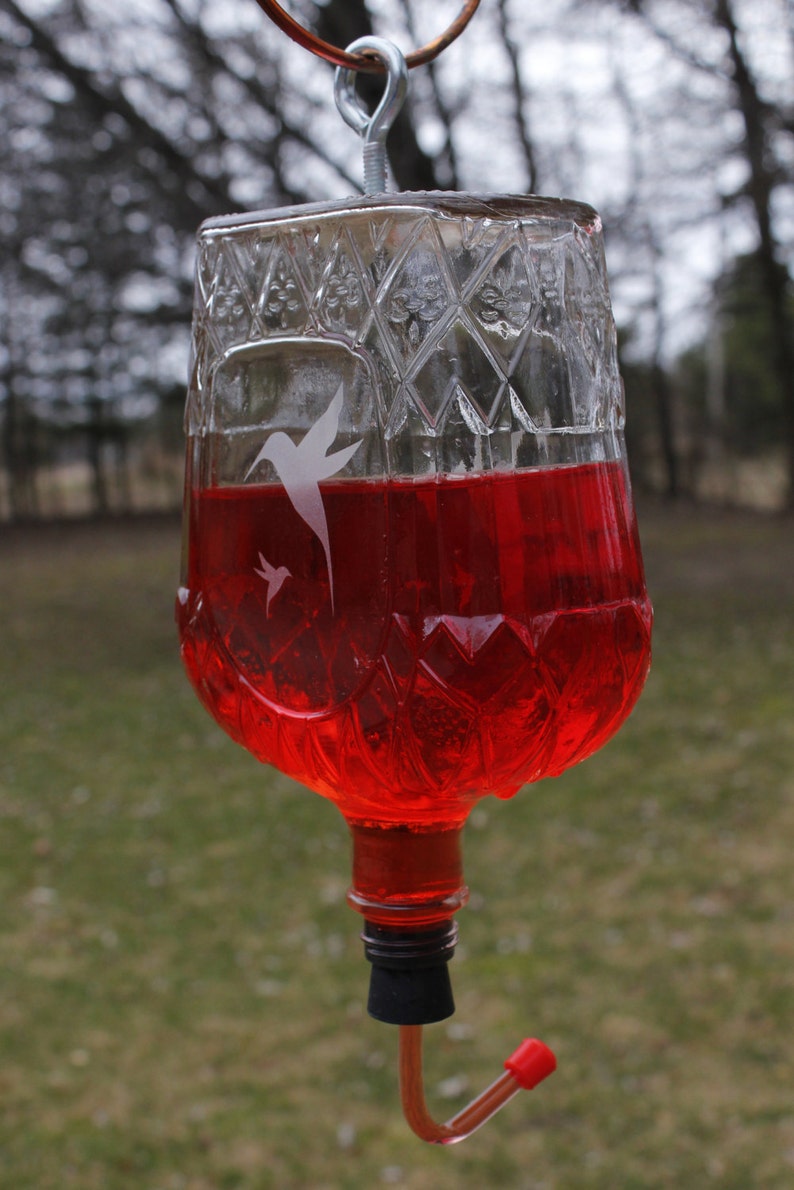 May include: A clear glass hummingbird feeder with a diamond-cut textured pattern filled with bright red liquid. The feeder features a white etched silhouette of two hummingbirds, a metal eye bolt at the top, and a curved feeding tube with a red tip.