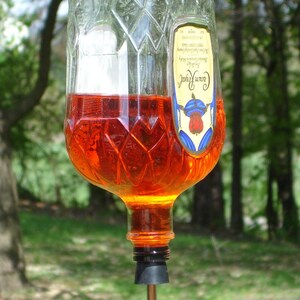 Op de afbeelding: Een omgekeerde Crown Royal glazen fles gevuld met oranje vloeistof, opgehangen aan een metalen oogbout. De fles heeft een ruitpatroon en een etiket. Een zwarte rubberen stop is in de hals geplaatst met een gebogen metalen buisje.