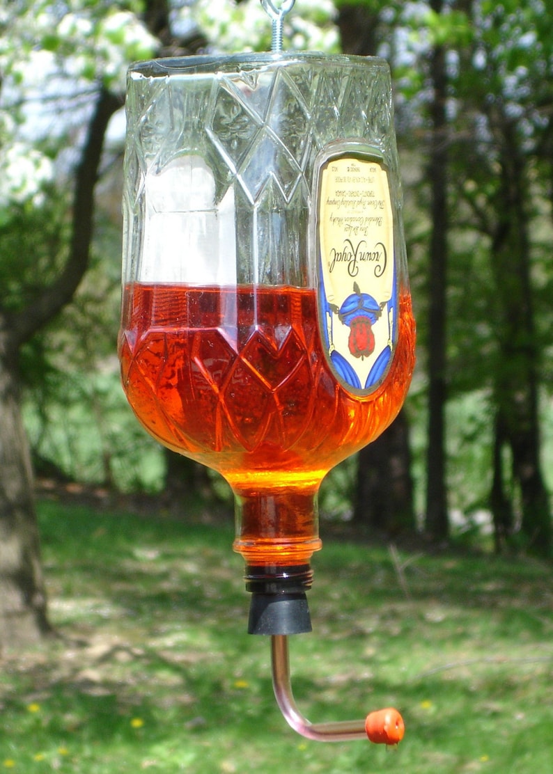 Op de afbeelding: Een omgekeerde Crown Royal glazen fles gevuld met oranje vloeistof, opgehangen aan een metalen oogbout. De fles heeft een ruitpatroon en een etiket. Een zwarte rubberen stop is in de hals geplaatst met een gebogen metalen buisje.