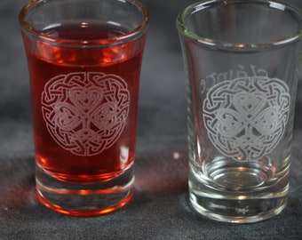 May include: Two clear glass shot glasses with an etched Celtic knot design featuring a shamrock. One glass is filled with a red liquid.