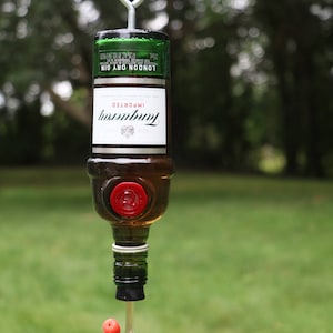 May include: A repurposed Tanqueray gin bottle transformed into a hummingbird feeder. The bottle is suspended by a metal hanger. The feeder has a red stopper and a clear, curved feeding tube. The background is a blurred green lawn.