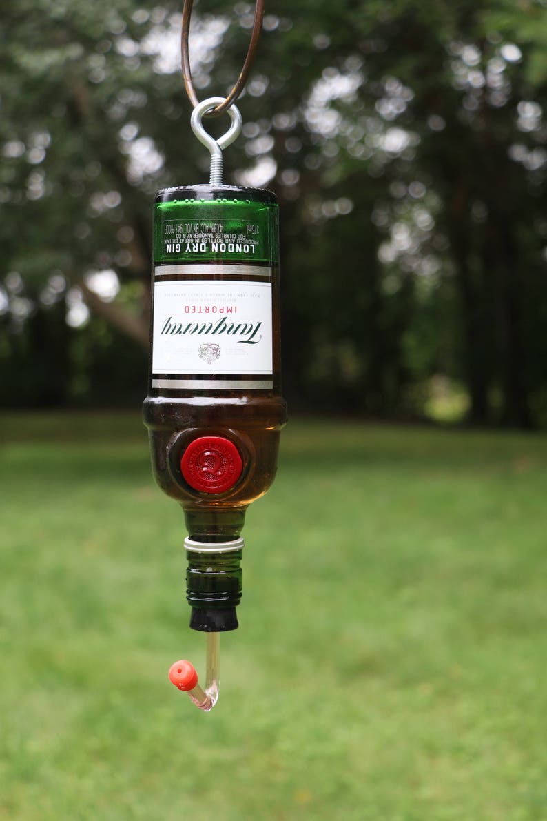 May include: A repurposed Tanqueray gin bottle transformed into a hummingbird feeder. The bottle is suspended by a metal hanger. The feeder has a red stopper and a clear, curved feeding tube. The background is a blurred green lawn.